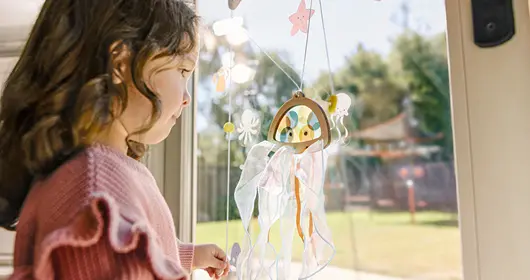 A young girl in a pink ruffled sweater gazes at the KiwiCo Ocean Quest project, which features a wooden jellyfish with flowing ribbon tentacles and colorful sea-life charms hanging in a sunlit window.